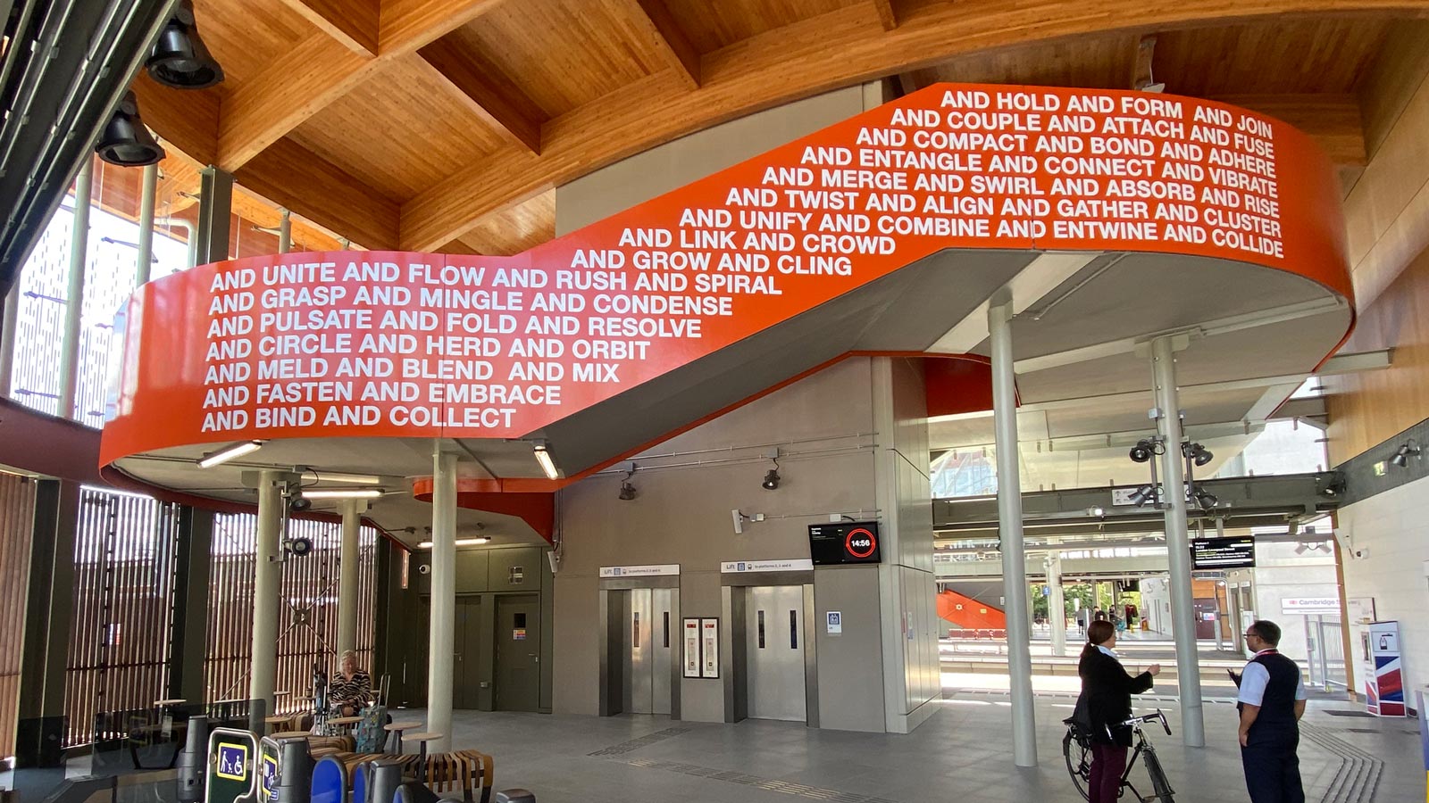 The east side of the Together We artwork in Cambridge South Station. The artwork in a strong orange colour with white caps lettering that says "And hold and form and join and couple and attach and fuse and compact and bond and adhere and entangle" etc.