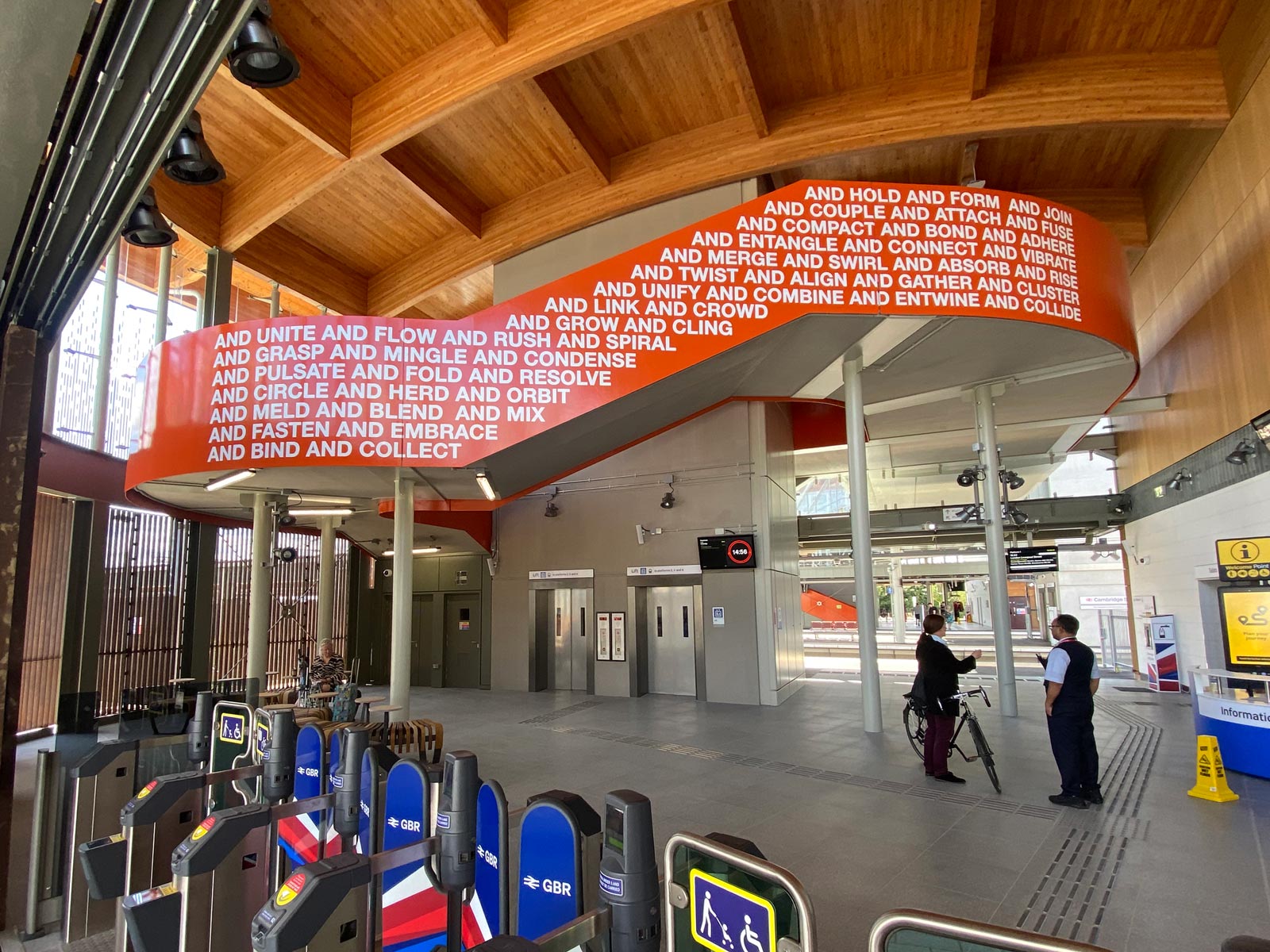 The east side of the Together We artwork in Cambridge South Station. The artwork in a strong orange colour with white caps lettering that says "And hold and form and join and couple and attach and fuse and compact and bond and adhere and entangle" etc.
