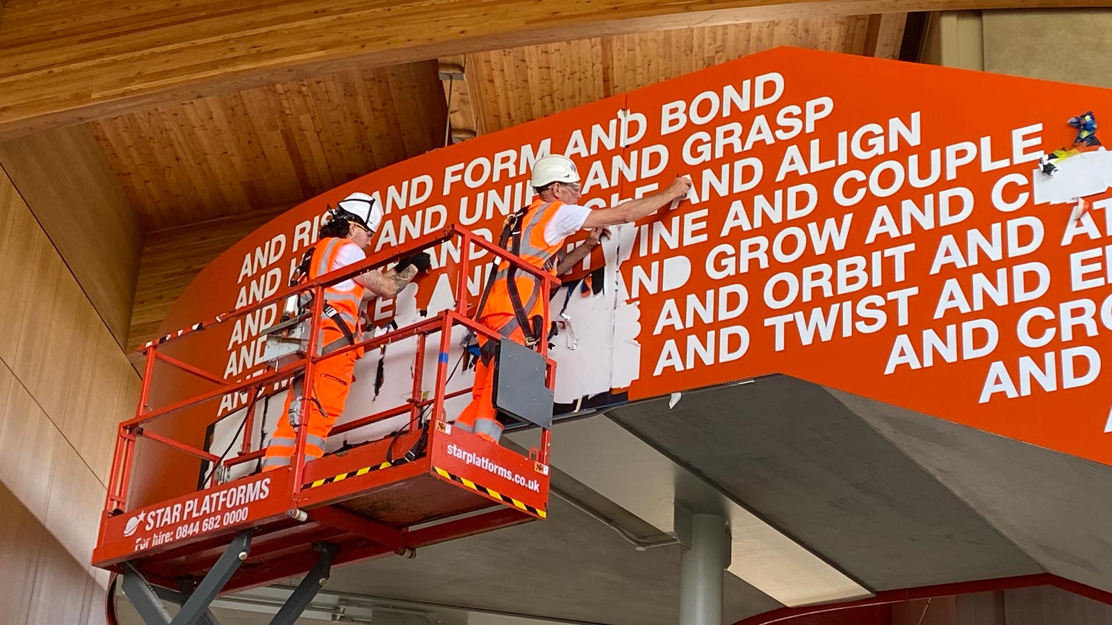 Two workers stand on a raised platform installing the Together We artwork in Cambridge South Station.