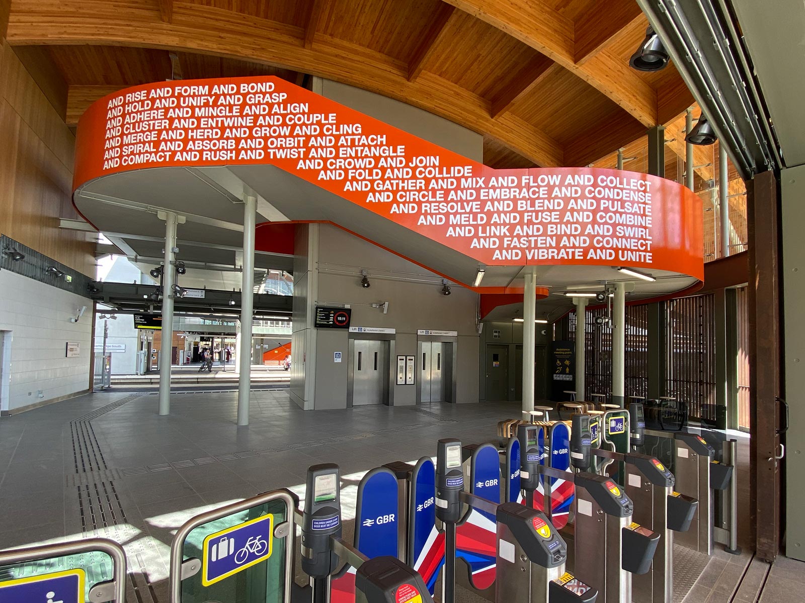 The west side of the Together We artwork in Cambridge South Station. The artwork in a strong orange colour with white caps lettering that says "and rise and form and bond and hold and unify and grasp and adhere and mingle and align" etc.