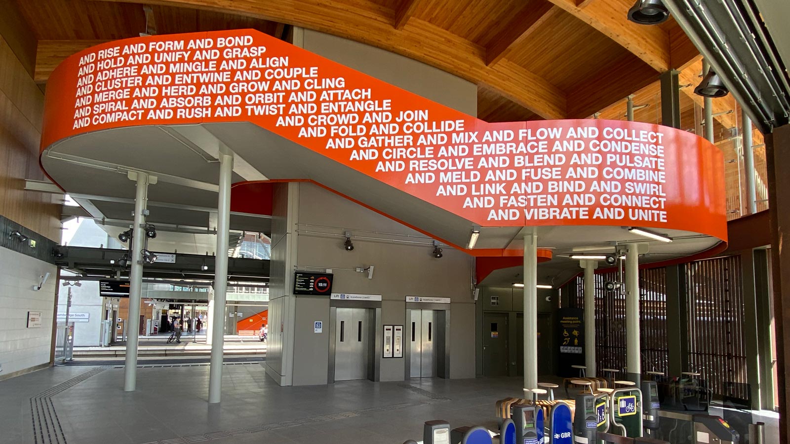 The west side of the Together We artwork in Cambridge South Station. The artwork in a strong orange colour with white caps lettering that says "and rise and form and bond and hold and unify and grasp and adhere and mingle and align" etc.