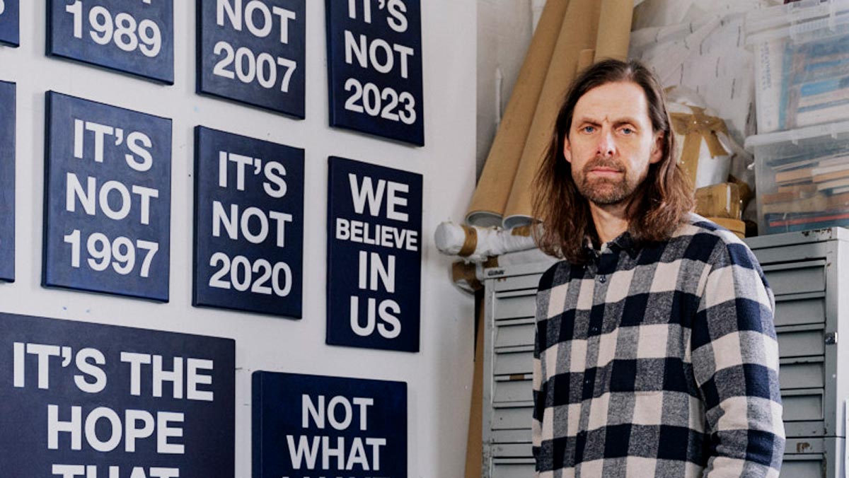 Artist Mark Titchner standing in his studio in front of several artworks with slogans such as "It's not 2020" and "We believe in us".
