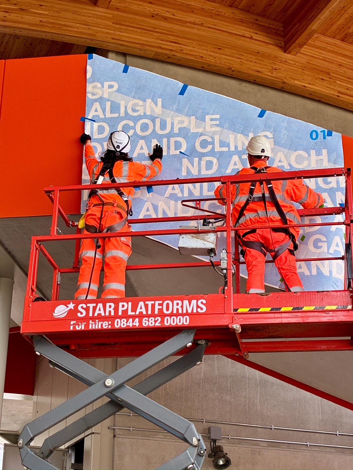 Two workers stand on a raised platform installing the Together We artwork in Cambridge South Station.